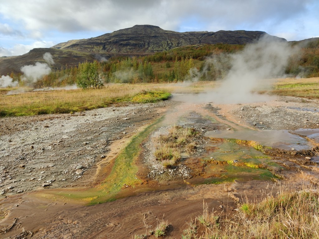 Kostea, höyryävä maisemakuva tämäkin. Taustalla paljas vuori. Kuvan tekee erilaiseksi värimaailma, jossa sekoittuvat harmaa sora, ruskea savi ja lähes neonvihreä sammal.