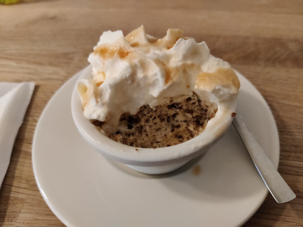 Kuvassa valkoinen lautanen, jonka päällä pieni kulho. Kulhossa erottuu ruskeankirjava leipämäinen täyte, jonka päälle on pursotettu kermavaahtoa ja vaaleaa kastiketta.