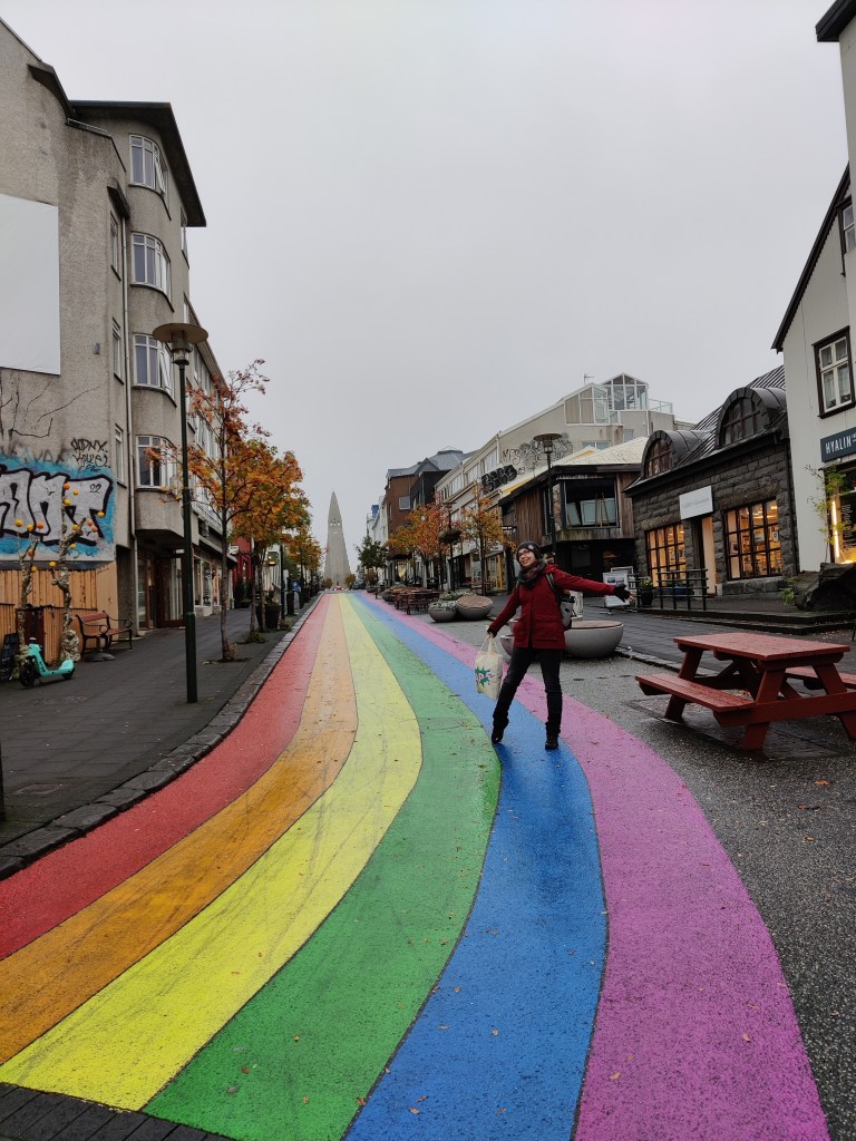 Kuvassa kävelykatu, johon on maalattu pitkä sateenkaatu. Sateenkaarikatu vie suoraan kohti Reykjavíkin maamerkkiä eli suurta kirkkoa. Katja poseeraa sateenkaarella kädet levällään.