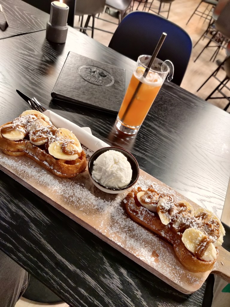 Kuvassa pöydällä puinen pitkä tarjoiluvati, johon on aseteltu kaksi banaanilla ja kinuskikastikkeella kuorrutettua peniksen mallista vohvelia. Näiden välissä on pyöreä kulho, jossa on vaniljajäätelöpallo. Koko komeus on koristeltu kookoshippusilla.