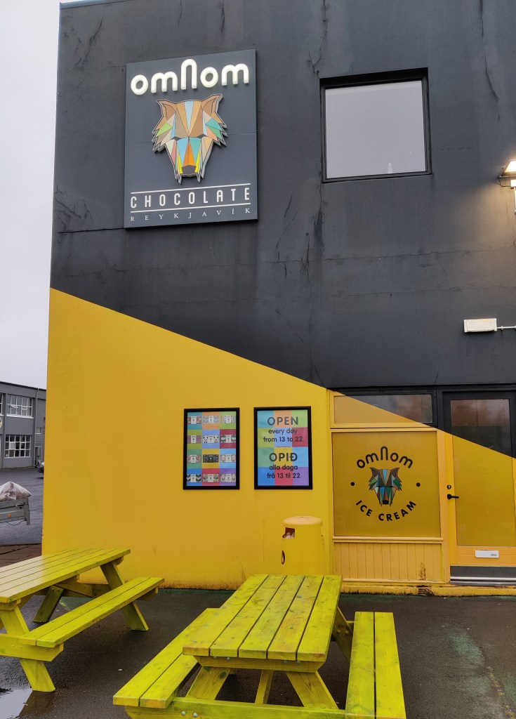 Om Nom -jäätelötehtaanmyymälän julkisivu, jonka edessä seisoo kaksi keltaista pöytäryhmää. Talon seinä on maalattu alhaalta keltaiseksi ja ylhäältä mustaksi. Sekä oven vieressä ikkunassa että korkealla kyltissä on logo omNom ja kuva kirjavasta, susimaisen hahmon päästä.