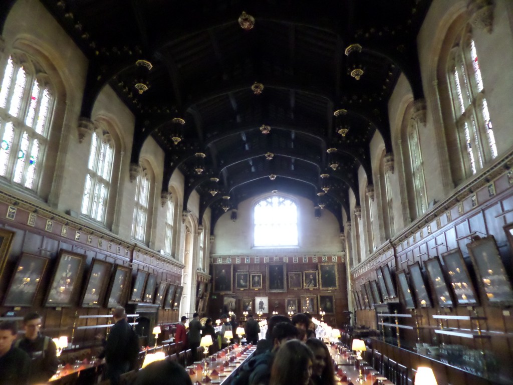 Suuri ruokasali, jossa pitkät pöydät, lasimaalausikkunat, paljon tauluja.