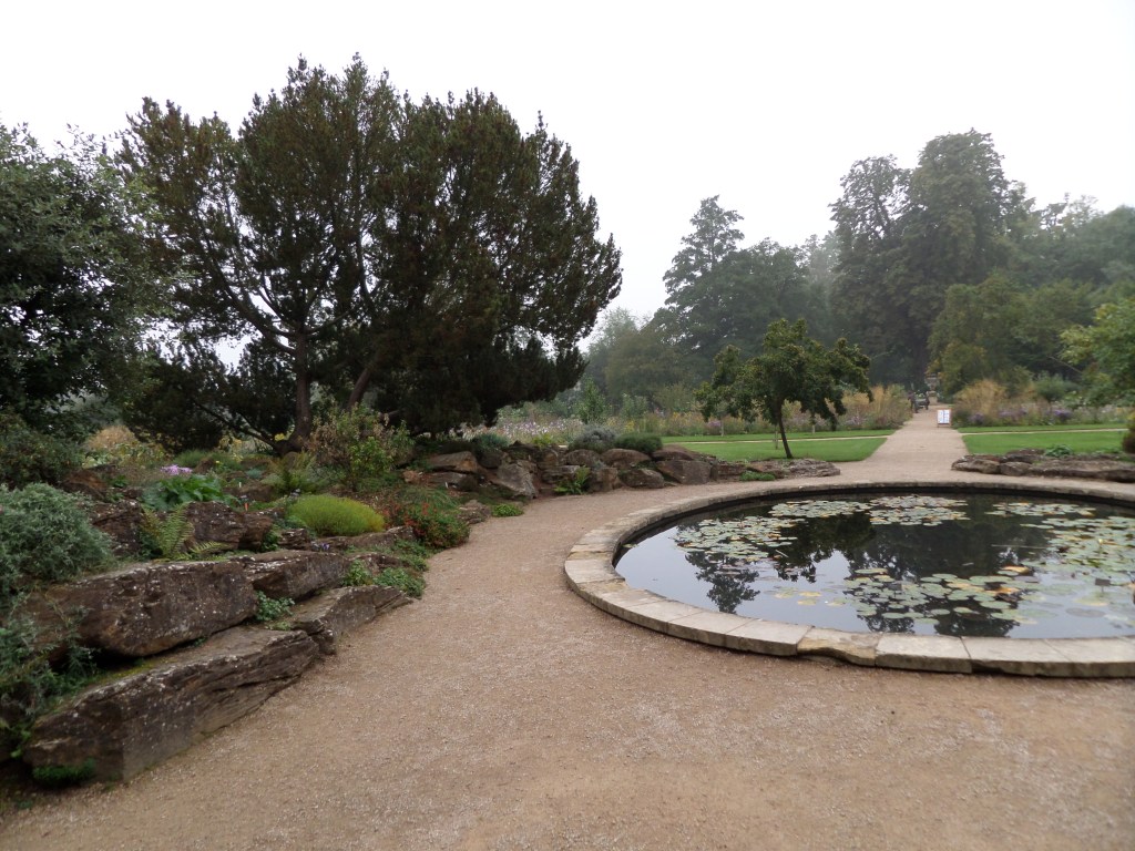 Kuva puutarhasta, jossa hiekalla päällystetty polku kiertää pyöreää vesiallasta. Taustalla puita.