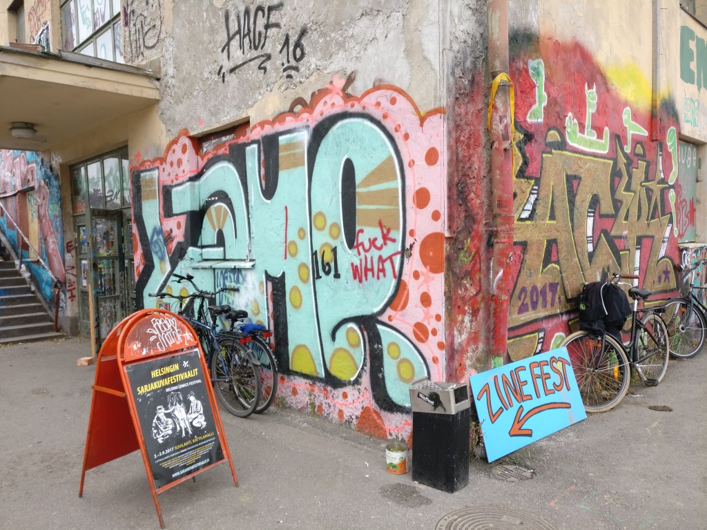Graffiteilla koristeltu seinä Helsingin sarjisfestareilla ja kyltti, joka ohjaa Zinefestin suuntaan.