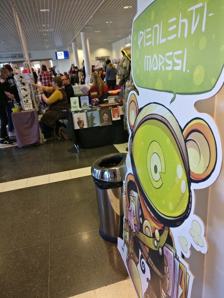 Lähikuvassa Pienlehtimarssin iso mainoskyltti, taustalla sarjakuva- ja taidemyyntikojuja Tampere Kupliissa.