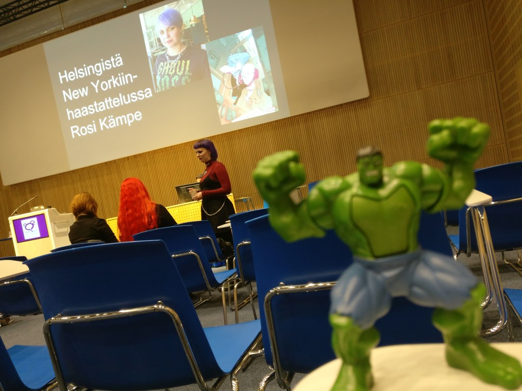 Etualalla sumea Hulk-figuuri, taustalla näkyy puhujanpöydän edessä sarjakuvataiteilija Rosi Kämpe aloittamassa omaa ohjelmaansa.