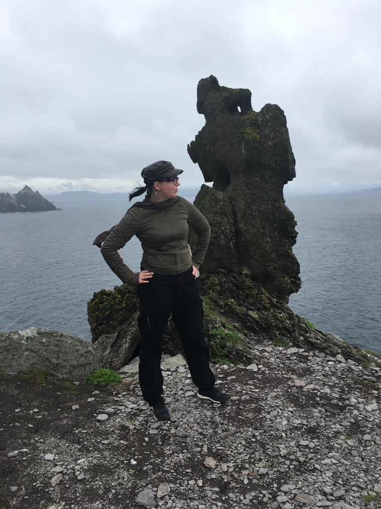 Kokovartalokuva Katjasta, joka poseeraa kivisaaren jyrkänteen reunalla olevan ison ja epämääräisen kiven edessä. Taustalla meri, pilvinen taivas ja toinen kalliosaari.