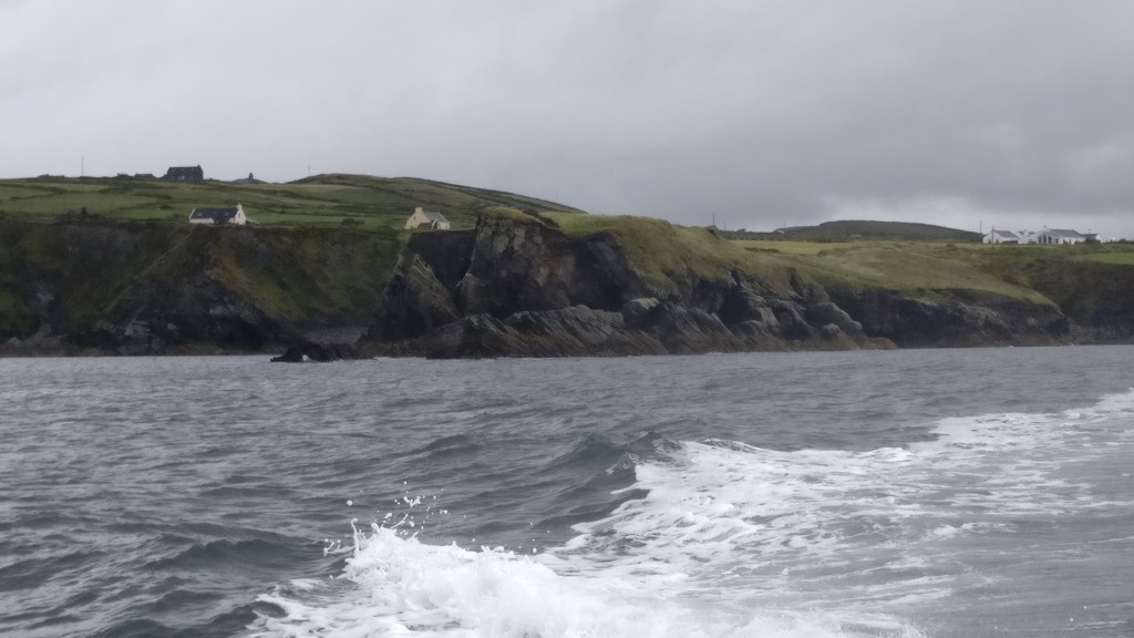 Kuva, joka on otettu veneestä. Etualalla merta, lähellä kallioinen ranta ja vihertävä, paljas maasto. Muutamia yksittäisiä taloja.