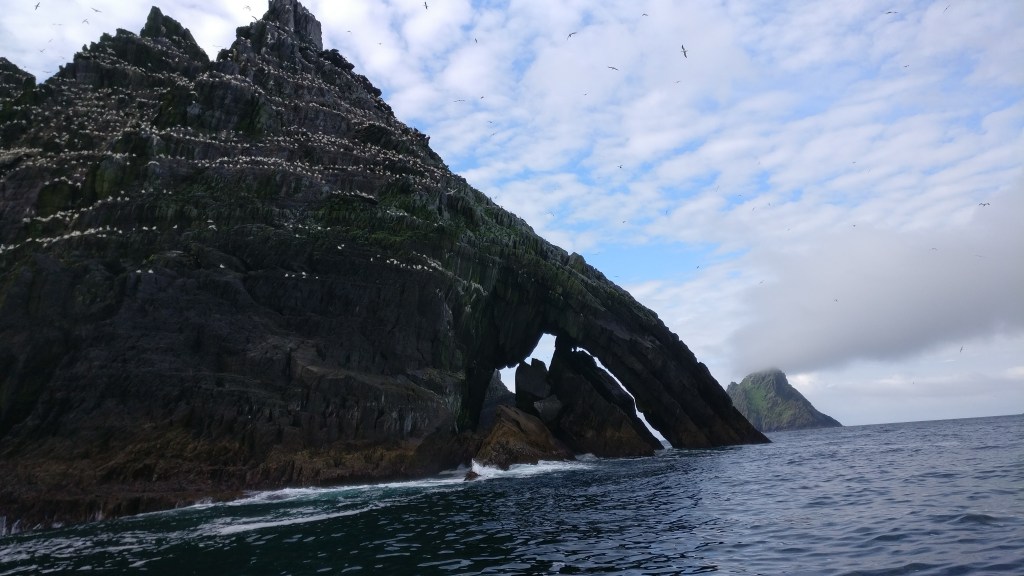 Kuvassa etualalla ylhäältä kapeneva tumma kalliosaari, jonka pinta on täynnä valkoisia täpliä, jotka ovat lintuja. Taustalla kauempana näkyy samanmallinen saari.