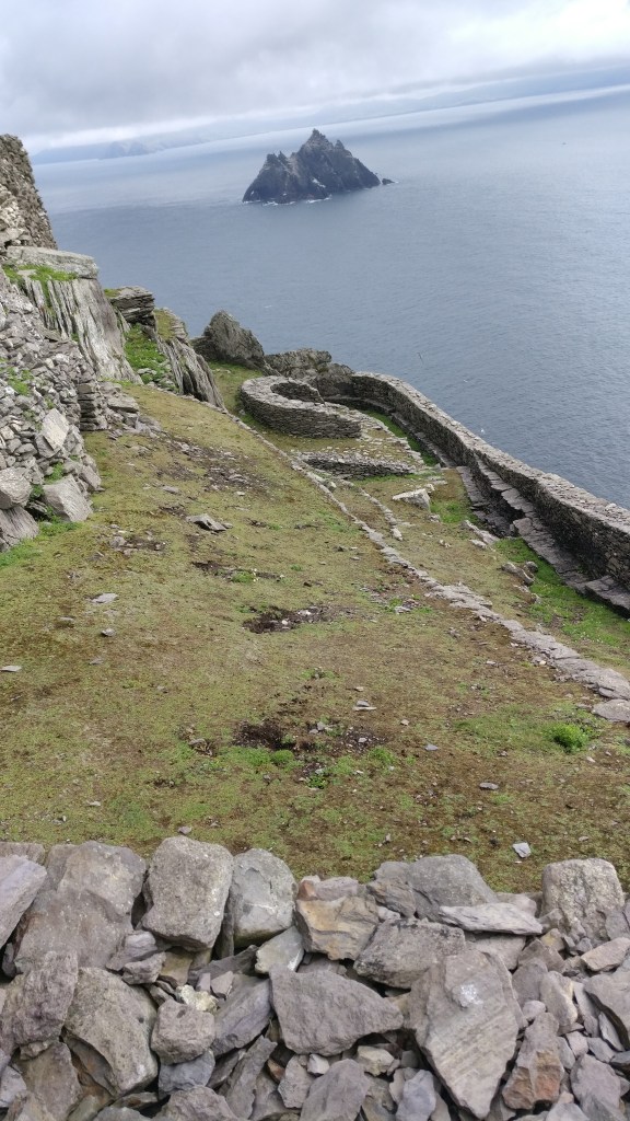 Kuva saaren huipulta, jossa on hieman nurmimaista kenttää ja kivistä kasattuja, mutkittelevia aitoja. Taustalla meri ja toinen kalliosaari.