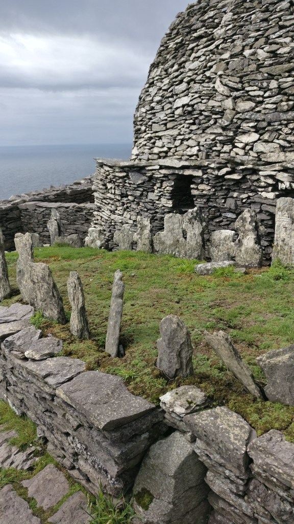 Kuva kivistä rakennetusta pyöreästä majasta, jonka eteen on pystytetty kiviä sammaleisiin maahan. "Pihaa" reunustaa matala kiviaita.