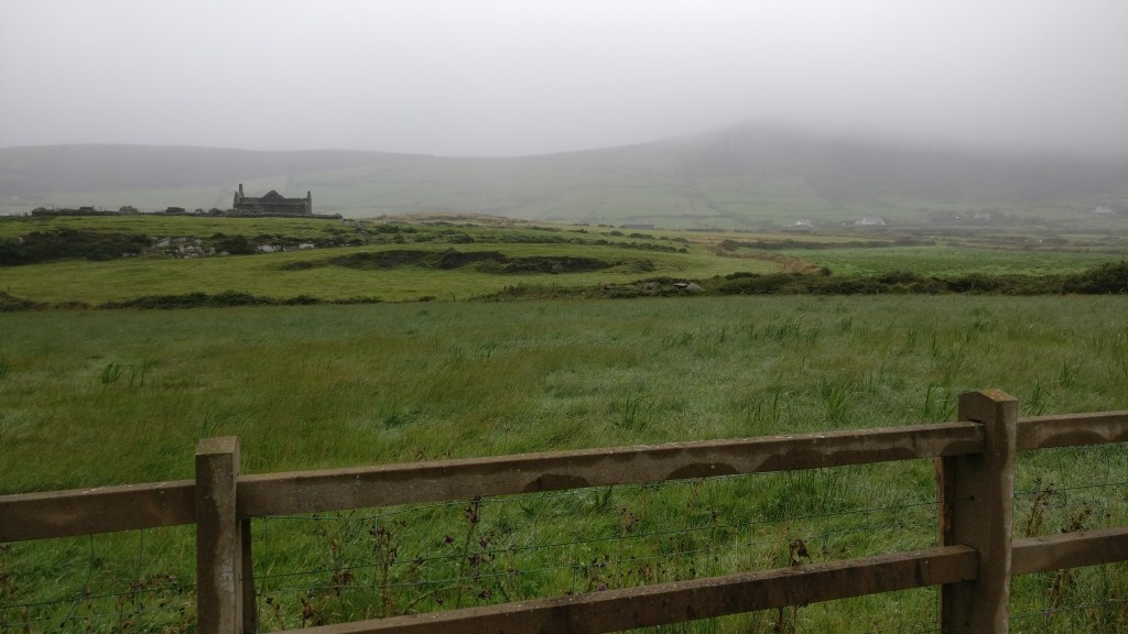 Maisema, jossa etualalla puinen aita, aidan takana niitty ja vanha rakennus. Taustalla kukkulat katoavat sumuun.