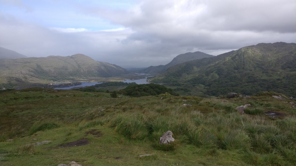 Maisemakuva, jossa etualalla karua niittyä, kauempana pensaikkoa. Maasto laskeutuu matalien vuorten keskellä olevalle järvelle.