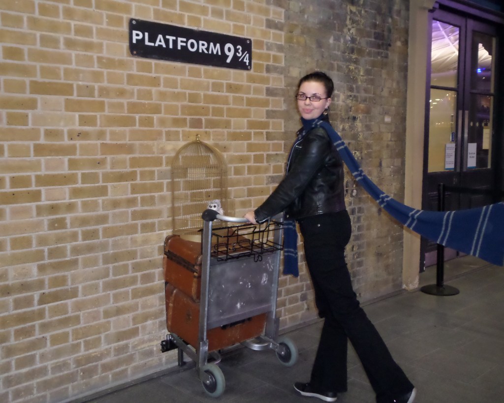 Kokokuva Katjasta mustassa nahkatakissa ja Korpinkynnen kaulaliinassa työntämässä kärryä seinän läpi Platform 9 3/4 -kyltin alla.