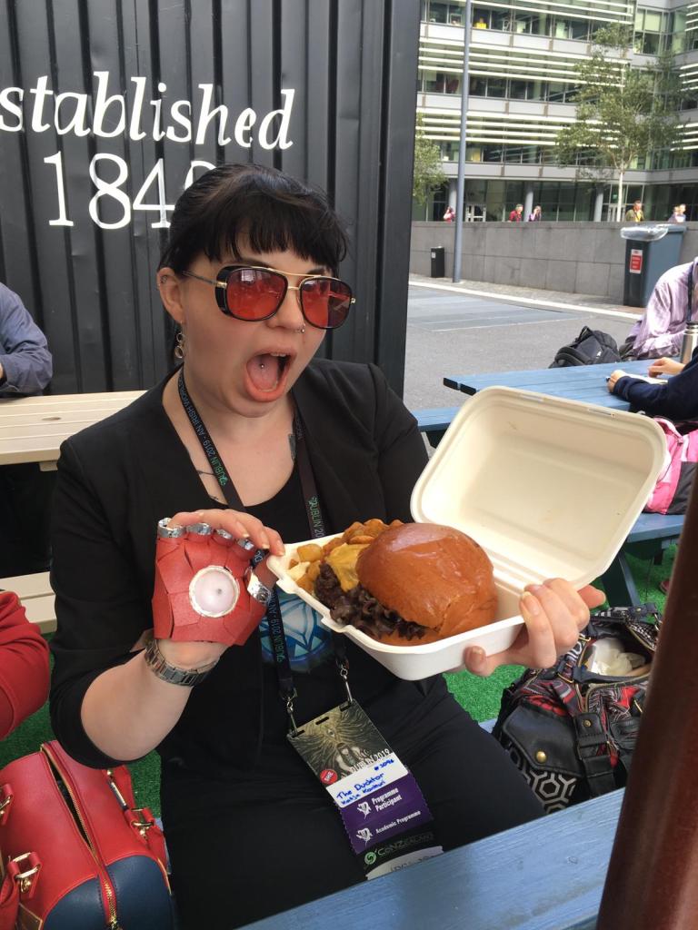 Kuva Katjasta, joka näyttää tuijottaa suu auki kameraa punaisten lasien takaa. Hänellä on käsissään kertakäyttörasia, jossa on iso hampurilaisannos.