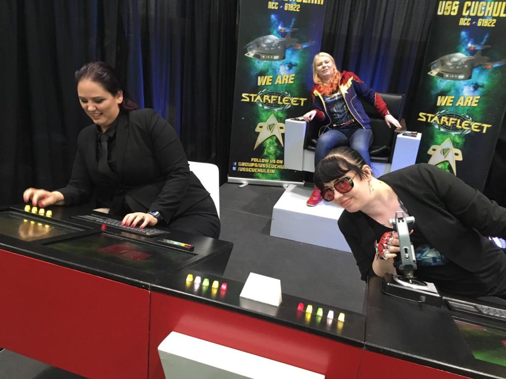 Ryhmäkuva Tupusta, Katjasta ja Tainasta, jotka istuvat Star Trek -aluksen "ohjaamossa": Tupu painelee nappuloita, Katjalla on jokin ohjain kädessään ja Taina istuu kauempana takana kapteenin penkillä. Kaikki ovat kallistuneet vasemmalle.