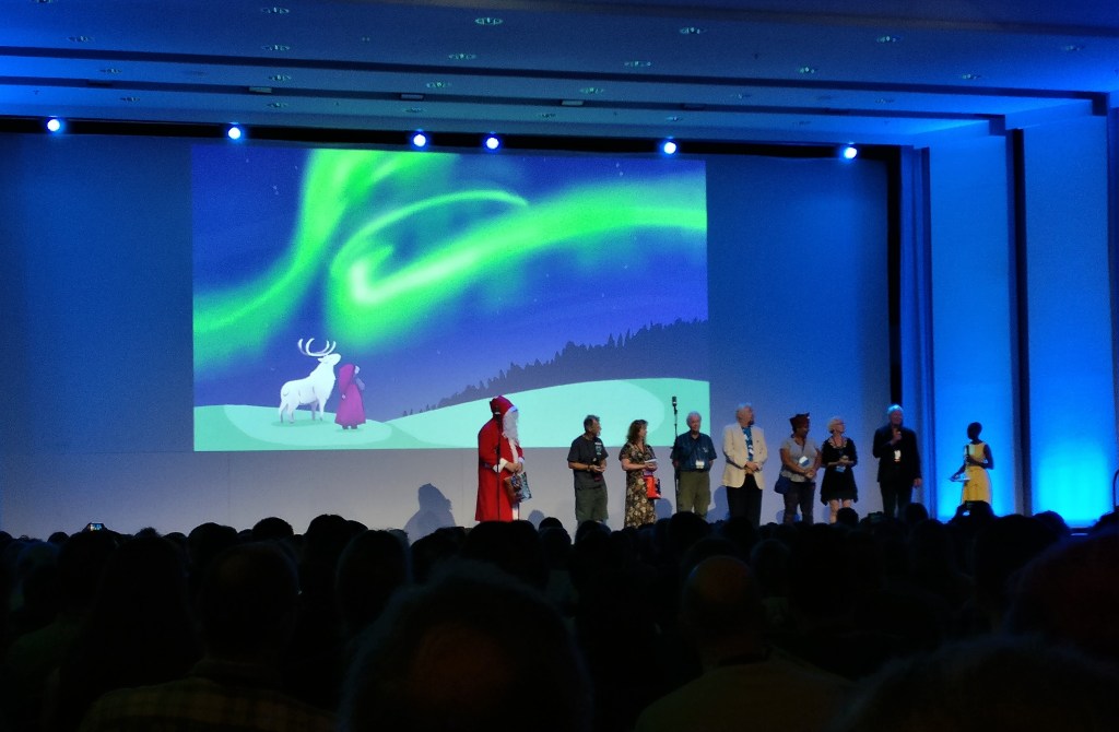 Kuva esiintymislavalta, jolla seisoo joukko Helsingin Worldconin kunniavieraita, juontaja ja Joulupukki. Taustalle on heijastettu talvinen piirroskuva, jossa on valkoinen poro, tonttu ja vihreitä revontulia.