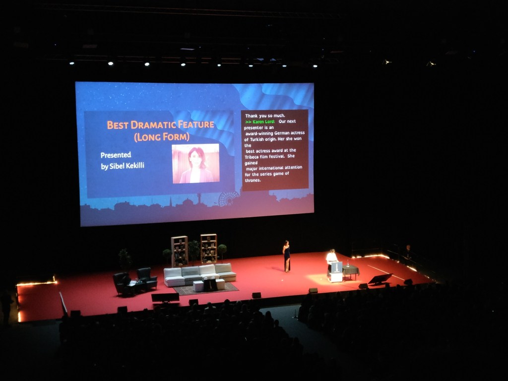Kaukaa parvekkeelta otettu kuva, jossa näkyy katsomon etummaiset rivit ja lava. Taustalle on heijastettu teksti "Best Dramatic Feature (Long Form) Presented by Sibel Kekilli" sekä hänen kuvansa. Lavalla seisoo juontaja ja mustaan mekkoon pukeunut SIbel Kekilli odottaa vuoroaan.