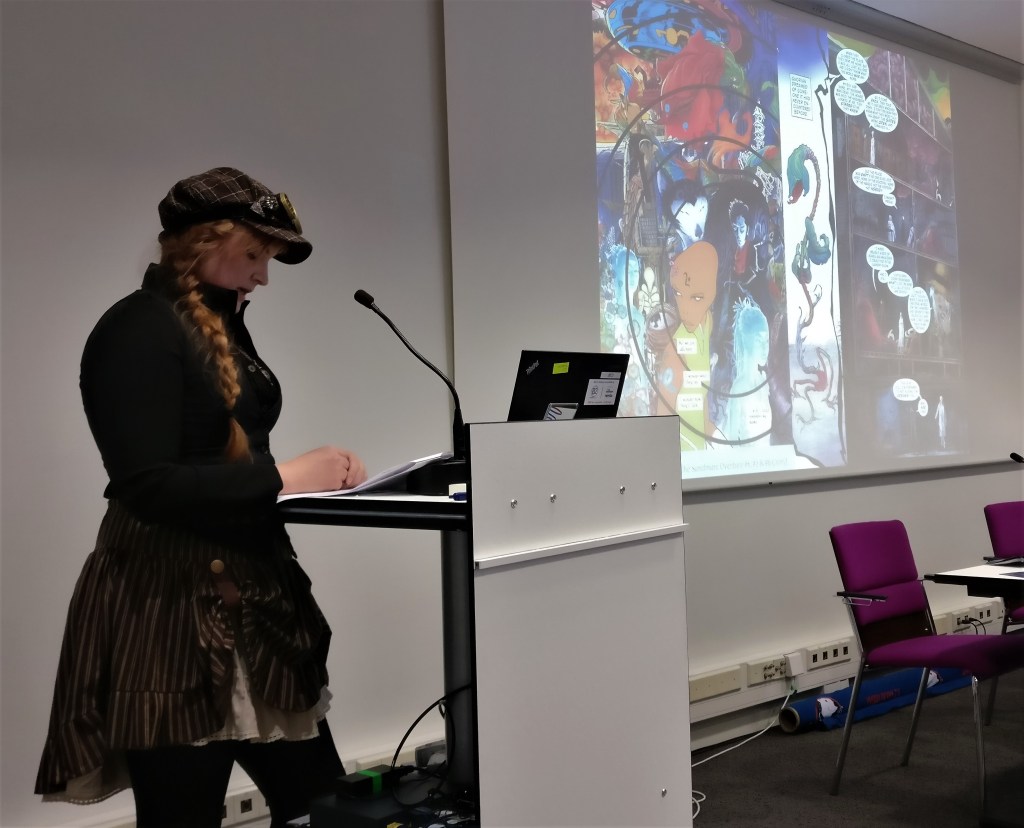 Kuva steampunk-henkisesti pukeutuneesta naisesta, jolla on vaalea pitkä tukka letillä, lätsä, jonka päällä vanhat "moottoripyöräilijän lasit" ja ruudullinen hame. Hän katsoo muistiinpanojaan ja puhuu mikrofoniin. Taustalla heijastettu kuva värikkäästä sarjakuva-aukeamasta.