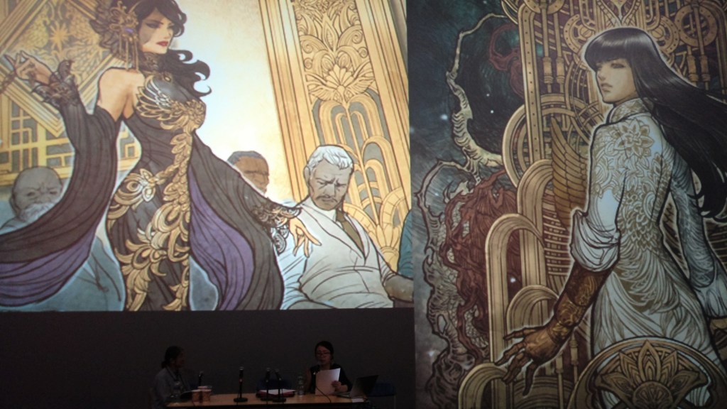 Kuva esiintyjistä, jotka näkyvät kovin tummina ja pieninä taustaa vasten: haastattelija ja taiteilija Sana Takeda. Huomion vie taustalle heijastettu Takedan sarjakuvateosten yksityiskohtainen kuvitus upeista, tummahiuksisista naisista ja koristeellisista taustoista.