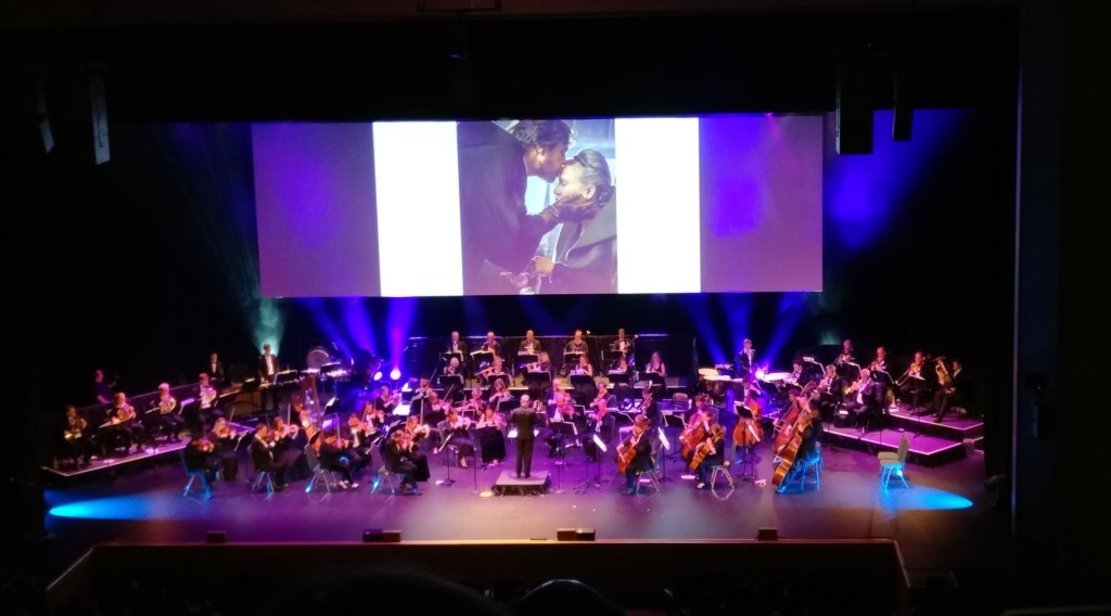 Kuva orkesterista lavalla soittamassa. Taustalla heijastettu valokuva uudesta Star Warsista, jossa Mark Hamill Luke Skywalkerina suutelee Carrie Fisherin kenraali Leia Organaa otsalle. 