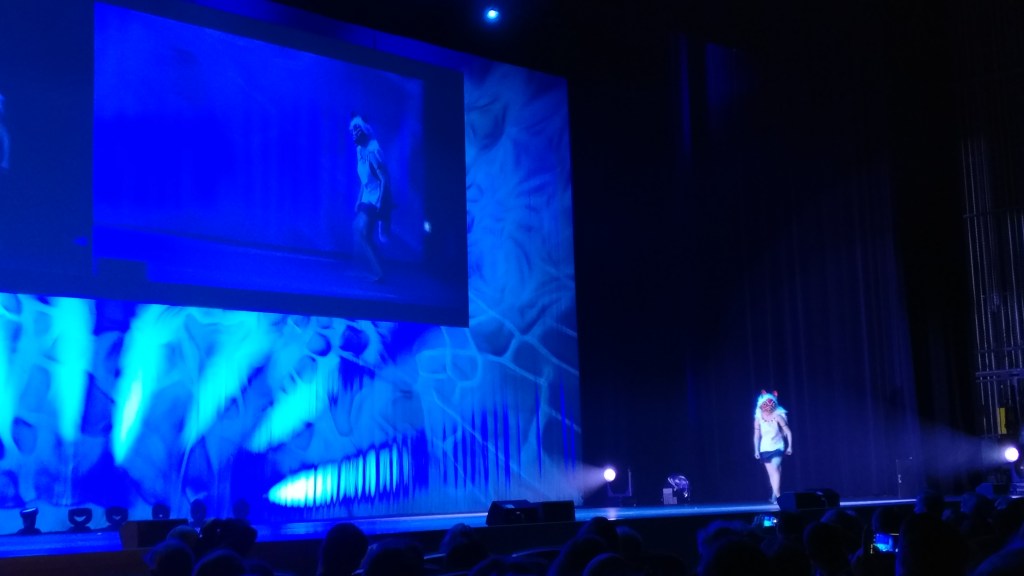 Kaukaa otettu kuva esiintymislavasta, jolla kävelee valkoiseen puseroon, naamariin ja peruukkiin pukeutunut hahmo. Kuva on sinertävä.