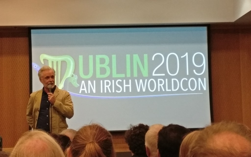 Kuva salista, jonka edessä seisoo harmaapäinen kirjailija mikrofonin kanssa. Taustalla heijastettuna teksti "Dublin 2019, an Irish Worldcon".