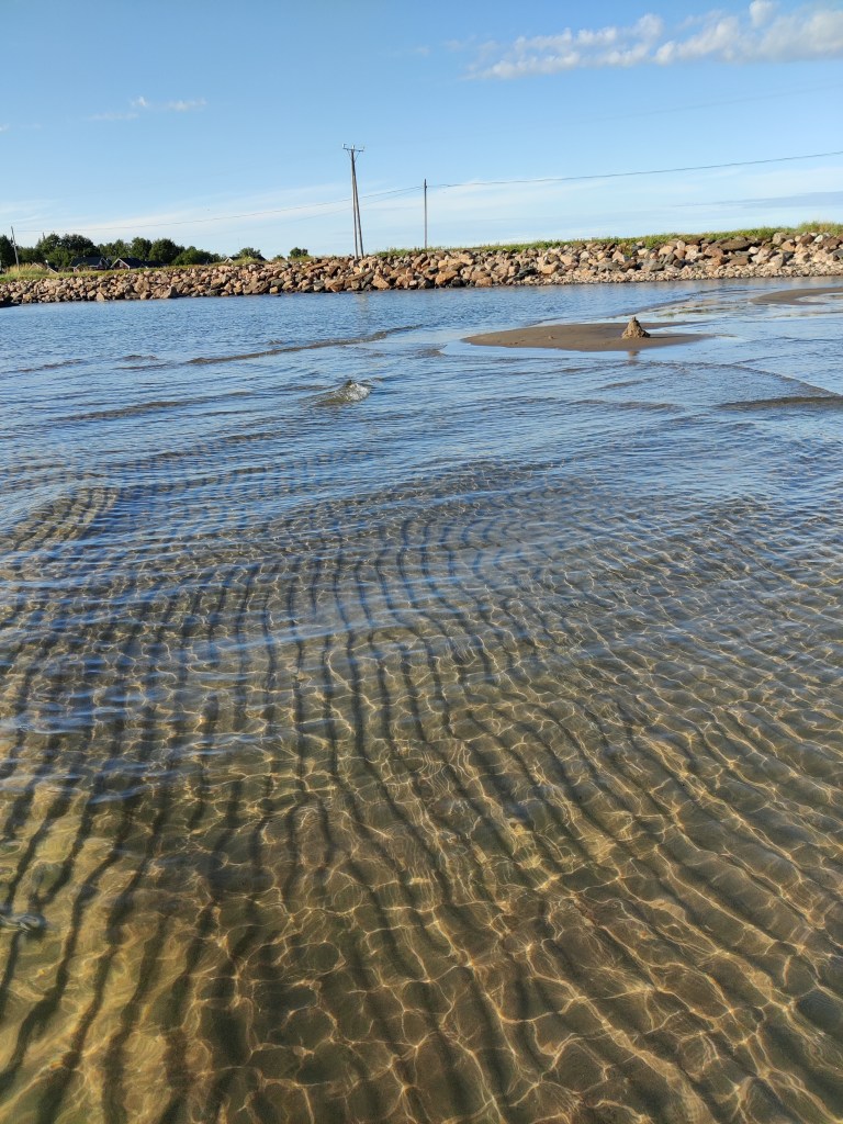 Vedestä käsin otettu kuva, jossa etualalla matala rantavesi, jonka pohjalle on muodostunut dyynejä. Kauempana hiekka kohoaa paikoin vedestä. Kaumpana näkyy kivien reunustama tie.