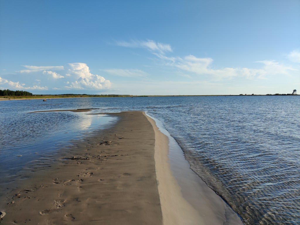 Merelle johtava veden keskellä nouseva hiekkadyyni, jossa erottuu jalanjälkiä. Taustalla näkyy puista maastoa.