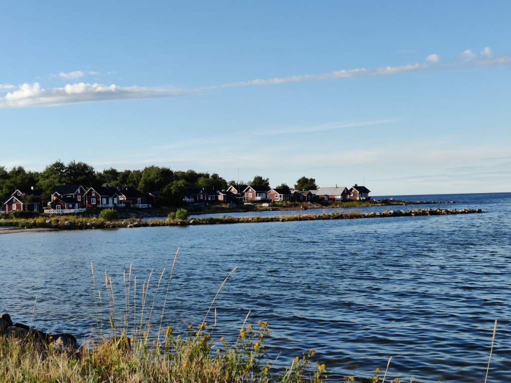 Maisemakuva mereen työntyvästä niemenkärjestä, jossa on idyllisiä punaisia pikkutaloja. Taivaalla muutama pilvi.