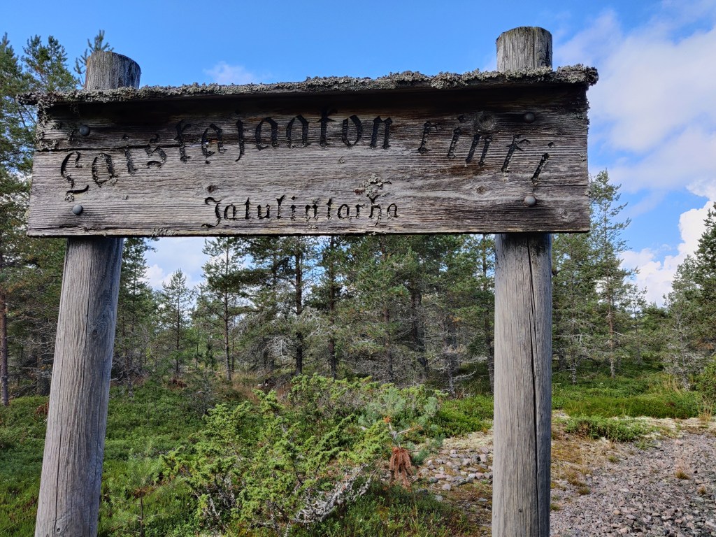 Kuva puisesta kyltistä keskellä havumetsää. Kyltissä lukee vanhoilla aakkosilla Laiskajaakon rinki, Jatulintarha.