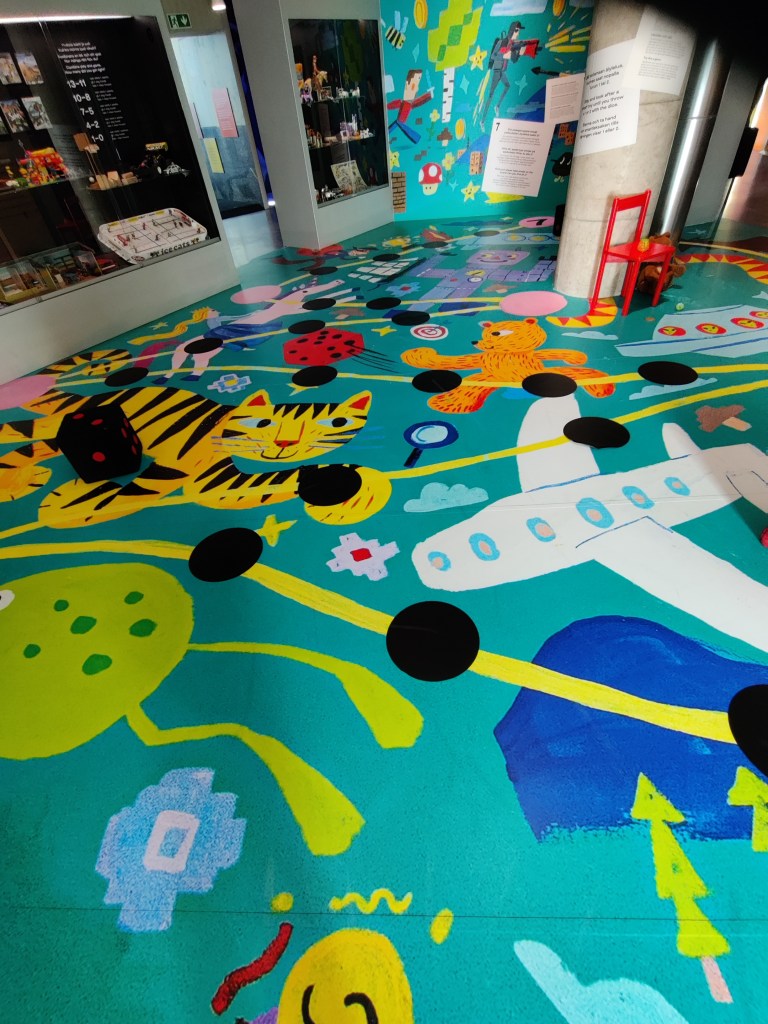 Kuvassa museon lattia, johon on maalattu koristeellisia eläimiä ja reitti, jossa on tietyin väliajoin mustia pylpyröitä. Lattialla on myös pehmeä noppatuoli. Kuvassa näkyy myös kaksi vitriiniä, joissa on leluja.