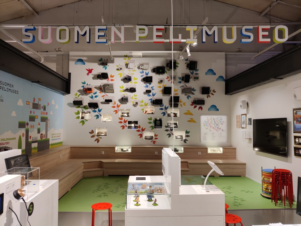 Kuvassa Pelimuseon sisätiloja: etualalla valkoisia vitriinejä, jotka esittelevät pelihahmoja. Taustalla leveä puinen istuskelualue. Takaseinällä esillä vanhoja pelikonsoleita, vasemmalle seinälle maalattu Super Marion kaksiulotteinen pelikenttä.