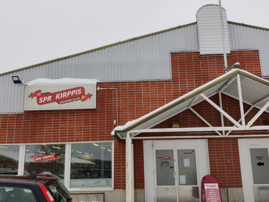 Kuva SPR-kirppiksen ulkoseinästä. Punatiilinen rakennus on teollisuushallimainen. Oven yläpuolella on harjakatos ja tervetuloa-kyltti. Seinässä on näkyvä valomainos, jossa lukee "SPR kirppis Kierrättää hyvää".