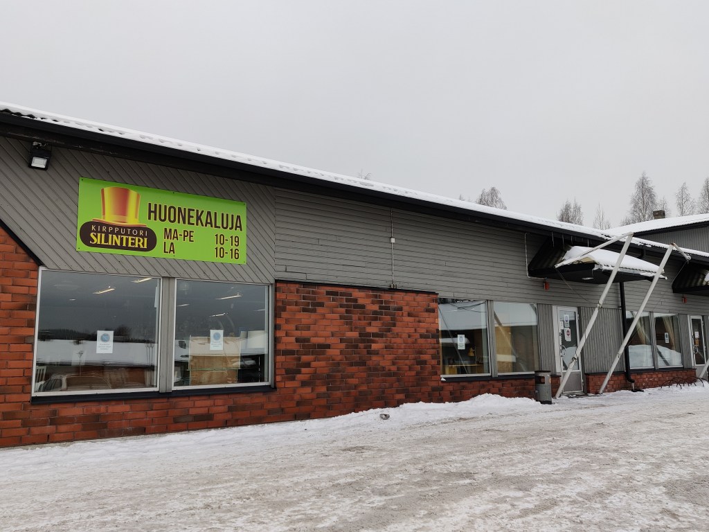 Kuva Kirpputori Silinterin ulkopuolelta. Tiilinen rakennus on matala ja tasakattoinen. Ikkunoiden yllä on neonvihreä mainoskyltti, jossa silinterin kuva ja liikkeen nimi. Kyltti mainostaa isolla HUONEKALUJA ja aukioloaikojaan.