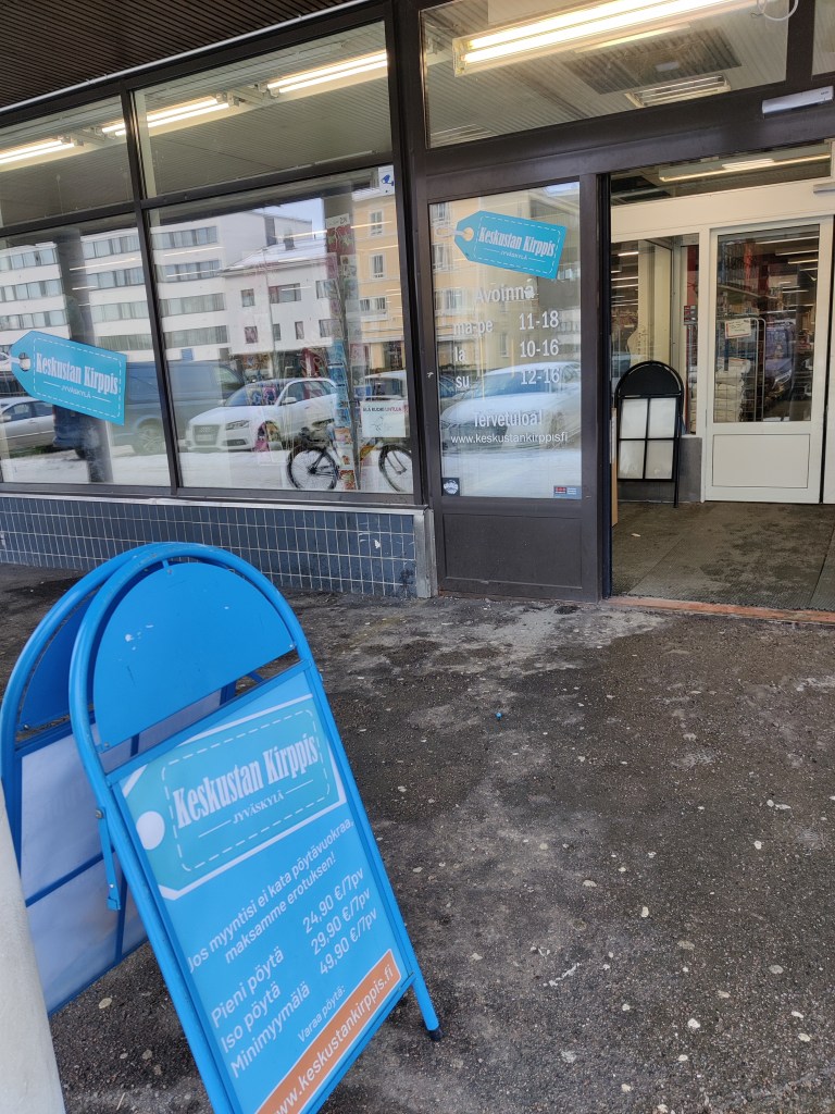 Kuva kirpputorin ulko-ovilta, joiden edessä on kirppiksen mainoskyltti pöytävuokrahintoineen.