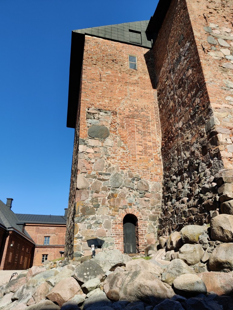 Kuva linnoituksen sisäpihalta, punatiilisen linnan ulkoseinästä, joka kohoaa korkeuksiin. Etualalla kivikkoa.