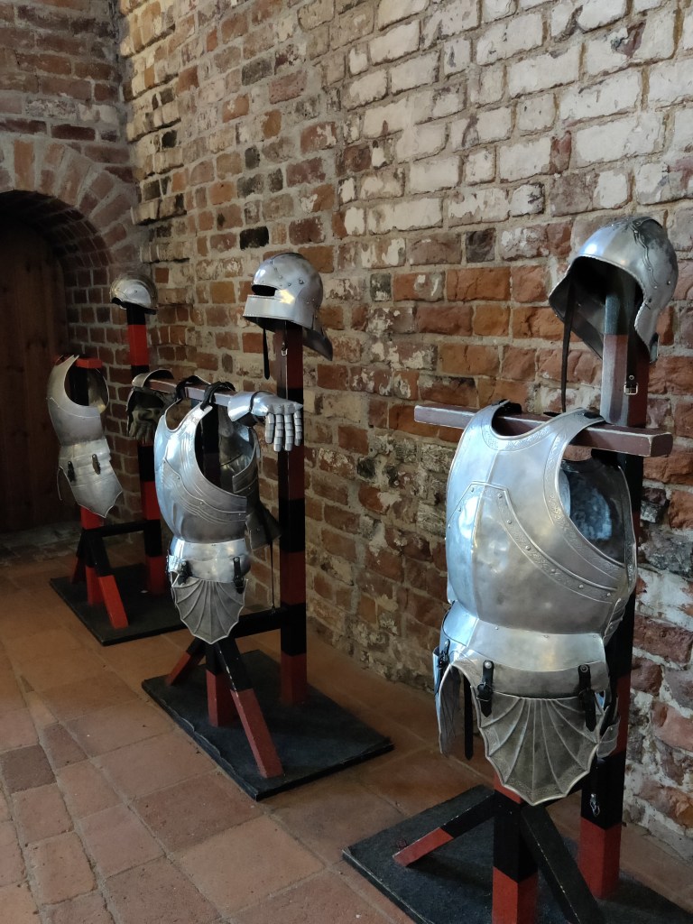 Kolme metallista haarniskan yläosaa sekä kypärät telineissä. Ne on tarkoitettu vieraiden kokeiltaviksi.