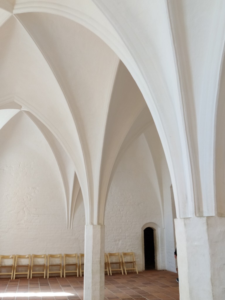 Kuva valkoiseksi maalatusta huoneesta, jossa valkoiset, reilusti ylöspäin levenevät pylväät kannattelevat kattoa. Lattia on ruskeaa laattaa. Seinän viereen on kasattu yksinkertaisia puisia tuoleja. Oikeassa nurkassa on mustana näkyvä oviaukko.