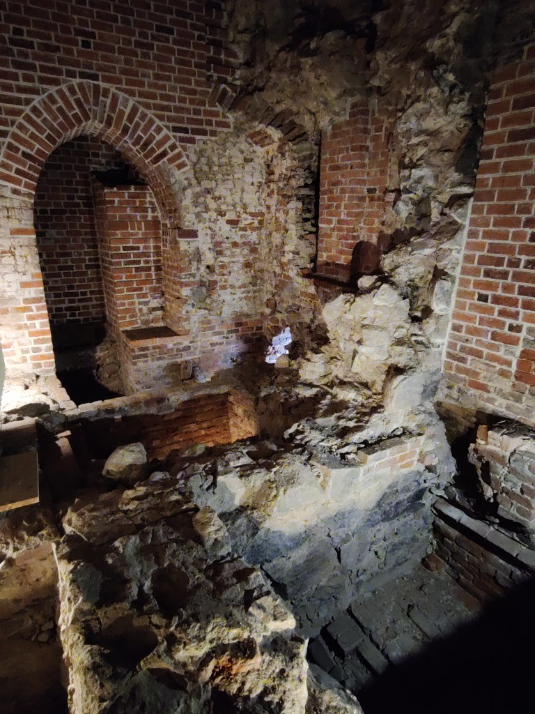 Kuva samasta rapistuneesta huoneesta sivustapäin otettuna. Pyöreä tiilistä rakennettu holvikaari on säilynyt ehjänä, mutta jokin iso rakennelma on murtunut keskellä huonetta. Kuva on otettu tasanteelta.