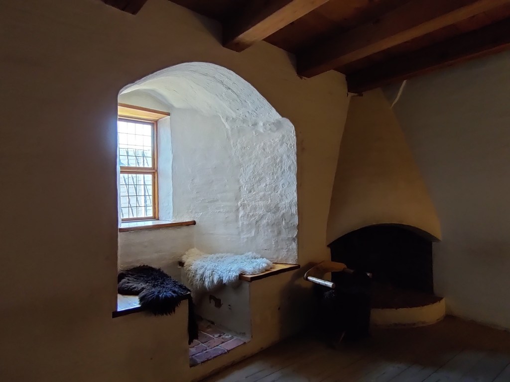 Kuvassa valkoisen huoneen takaseinä, jossa iso ikkunasyvennys penkkeineen. Penkkien päällä on eläinten taljoja. Oikealla puolella huoneen nurkassa on takka. Katto on tummanruskea parrukatto.