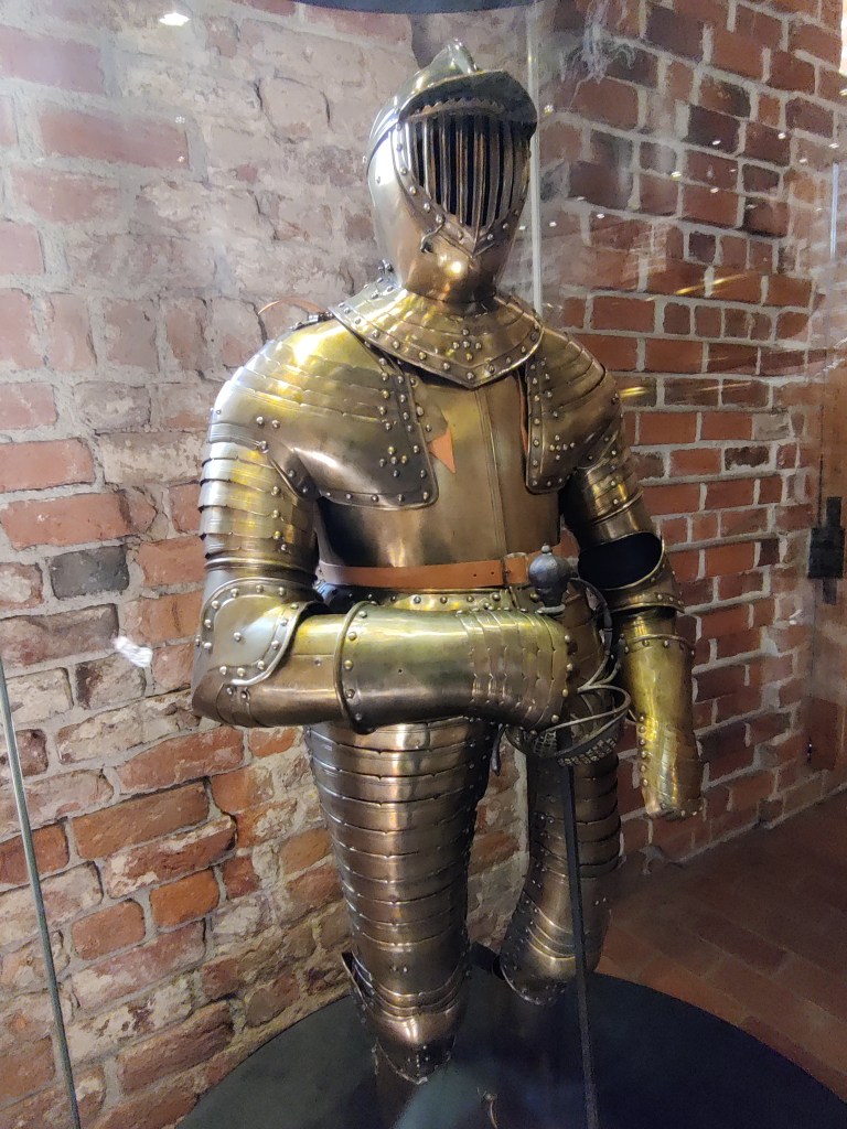 Kuvassa kultaisen värinen täyshaarniska,  jonka kypärässä on silmikko. Haarniskan oikea käsi nojaa pistomiekkaan.