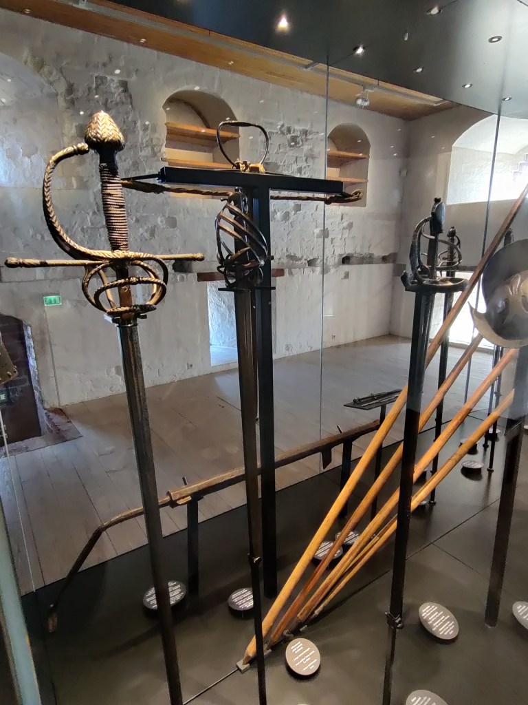 Kuvassa useita aseita. Etualalla kolme erilaisella kahvalla varustettua pistomiekkaa. Kauempana näkyy puisia keihäitä ja osa harmaasta kypärästä.