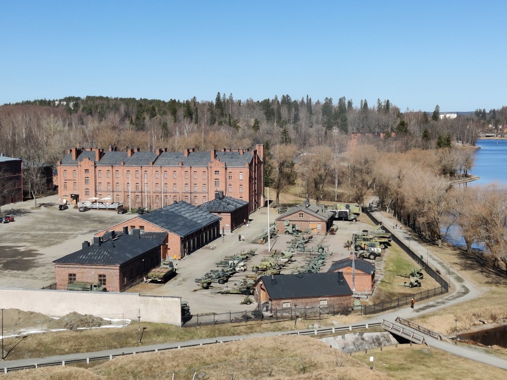 Ylhäällä otettu kuva, jossa näkuu valtava punatiilinen rakennus ison pihan takana. Piha jakaantuu kahtia: vasemmalla puolella tyhjä hiekkakenttä, oikealla punatiilisten pienempien rakennusten ja aidan rajaama alue, jossa näkyy tummanvihreitä tankkeja ja tykkejä.