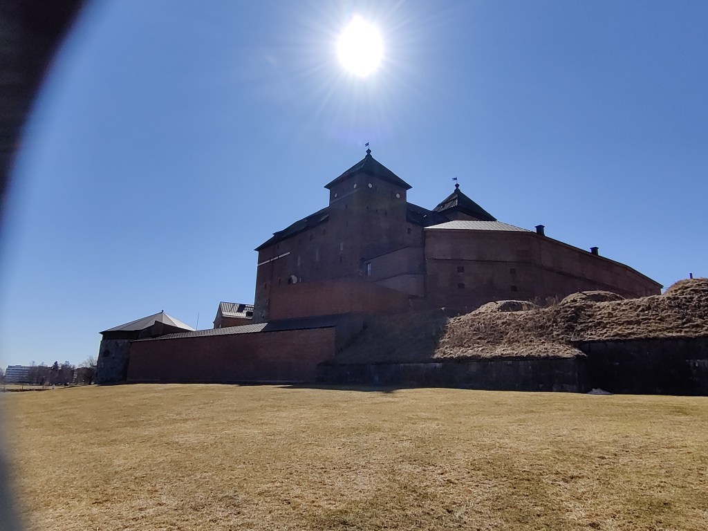 Kuva Hämeen linnasta ulkomuurien toiselta puolelta otettuna. Linna kohoaa suoraan edessä punaisena ja tyylikkäänä. Aurinko paistaa suoraan kameraan siniseltä taivaalta.