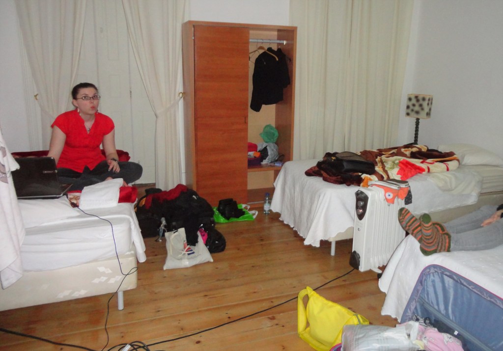 Kuva hotellihuoneesta, jossa on vaaleat seinät ja valkoiset verhot. Huoneessa näkyy kolme sänkyä ja ruskea vaatekaappi. Sängyillä on tavarakaaos. Punapaitainen Katja istuu vasemmanpuolimmaisella sängyllä läppärin kanssa ja irvistelee kuvaajalle. Siskon villasukkaiset jalat näkyvät oikeassa reunassa.