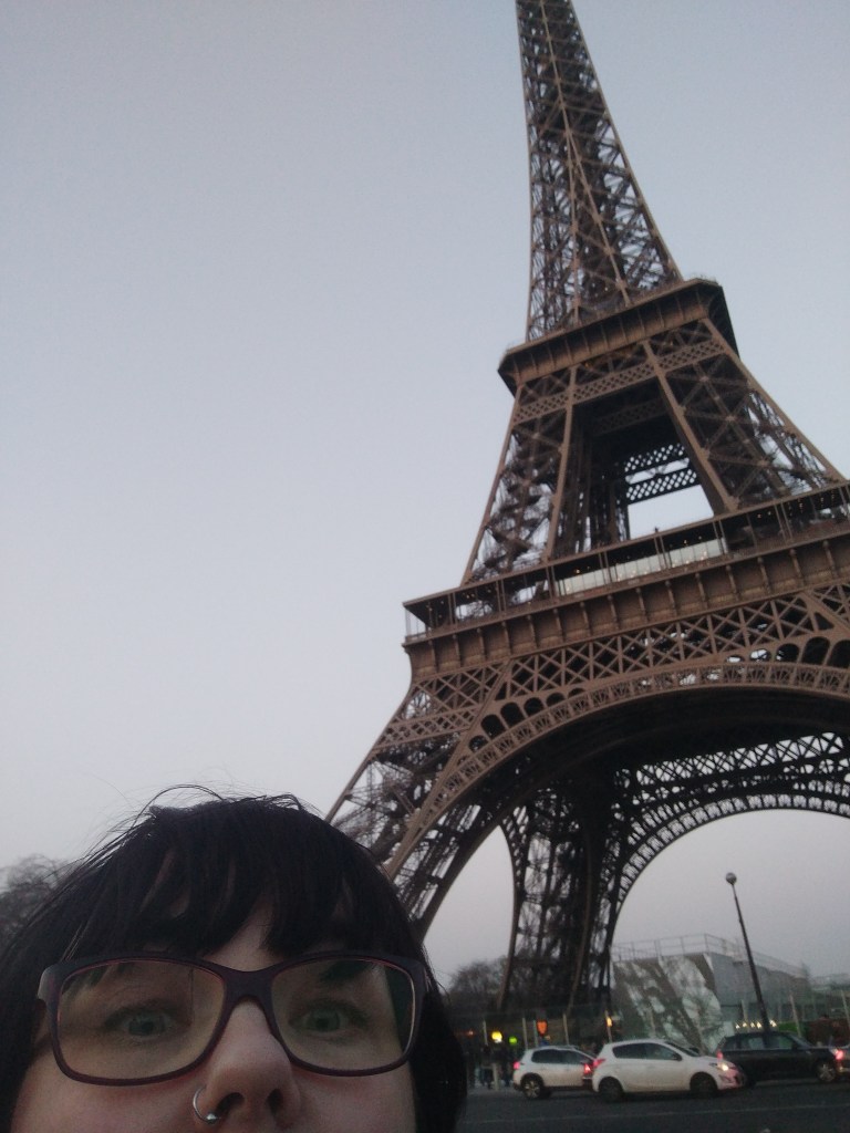 Erikoislähikuvaselfie, jossa Katjasta näkyy nenä ja silmät. Taustalla kuvaa hallitsee Eiffelin torni. Taivas on pilvinen ja harmaa.