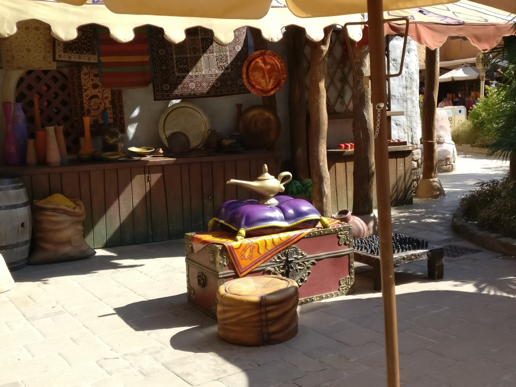 Kuvassa basaarimainen katos, jossa esillä erilaisia esineitä ja mattoja. Katoksen alla pieni kirstu, jonka päällä on violetti tyyny. Ja tyynyn päällä on Aladdinin taikalamppu.