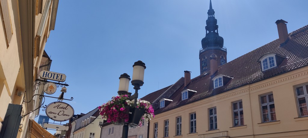 Kuva talojen yläosista: vasemmalla näkyy kyltti, jossa lukee isolla HOTEL ja pienemmällä Hotel am Dom. Sen vieressä on kaksiosainen lyhtypylväs, jossa roikkuu kukkaruukkuja. Oikealla rakennusten takana näkyy kirkontorni.