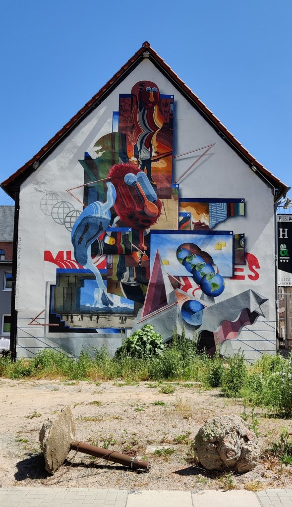 Kuva harjakattoisen talon päädystä, johon on maalattu värikäs seinämaalaus. Kuvassa on useita kuvia päällekkäin ja lomittain, eikä siitä oikein saa selvää, mitä se esittää.